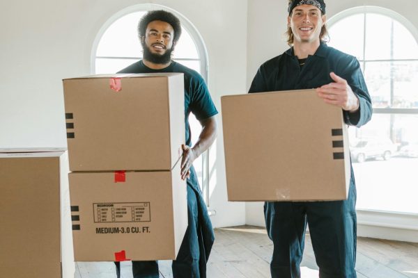 Two smiling movers carrying cardboard boxes in a bright, airy room with wooden floors.